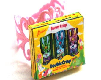 May include: A miniature box of Bunny Crisp chocolate bars with a yellow background and a picture of three bunnies in green, blue, and purple foil wrappers. The box says "Bunny Crisp" and "Double Crisp" on it. The box is sitting on a pink plastic ring.