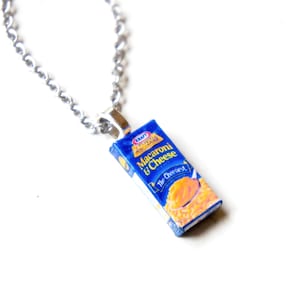 May include: A silver chain necklace with a charm that looks like a box of Kraft Macaroni & Cheese. The box is blue and yellow and has the text "Kraft Macaroni & Cheese" and "The Cheesiest."