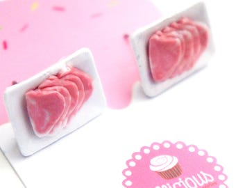 May include: A pair of novelty earrings featuring miniature pink meat slices arranged on white trays. The earrings are set against a pink background with a logo that says "Jillicious" and a cupcake illustration.