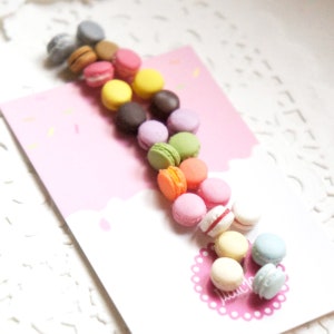 May include: A collection of colorful macarons arranged in a line on a pink background. The macarons are in various shades of pink, yellow, green, blue, and orange.