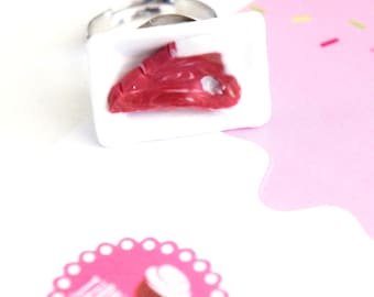 May include: A novelty ring featuring a miniature piece of raw red meat, presented on a white tray, set against a pink background. The ring's band is silver-toned. The overall theme is food-related jewelry.