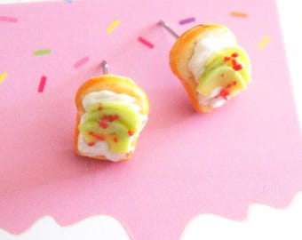 May include: A pair of miniature food-themed stud earrings. Each earring is shaped like a slice of toast topped with white cream, kiwi slices, and red sprinkles. The earrings are set against a pink background with colorful sprinkles.