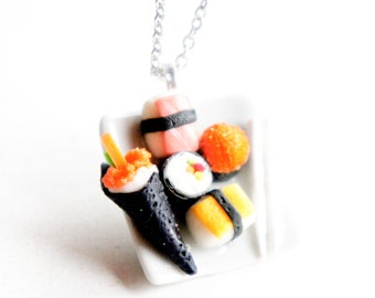 May include: A silver chain necklace with a white plate charm featuring a variety of sushi made from polymer clay. The sushi includes a maki roll, nigiri, and a hand roll.