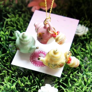 Könnte beinhalten: Eine goldene Kettenkette mit drei Porzellan-Teekannen und Cupcakes als Anhänger. Die Teekannen sind mit floralen Mustern in Rosa, Grün und Gelb verziert. Die Cupcakes sind mit rosa Zuckerguss und Streuseln verziert.