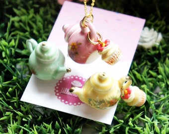 Puede incluir: Un collar de cadena dorada con tres teteras de porcelana y cupcakes como colgantes. Las teteras están decoradas con motivos florales en rosa, verde y amarillo. Los cupcakes están cubiertos con glaseado rosa y chispas.