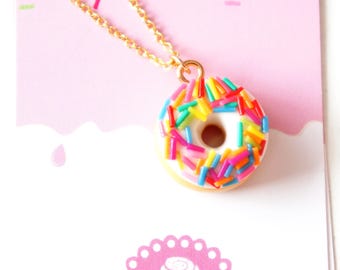 Könnte beinhalten: Eine goldene Halskette mit einem weißen Donut-Anhänger, der mit bunten Streuseln bedeckt ist.