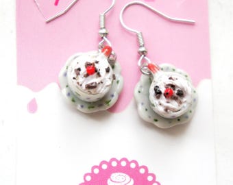 May include: A pair of whimsical cupcake earrings. Each earring features a miniature cupcake with white frosting, chocolate sprinkles, and a red cherry on top, set on a small, scalloped plate. The earrings hang from silver-colored fish hook ear wires.