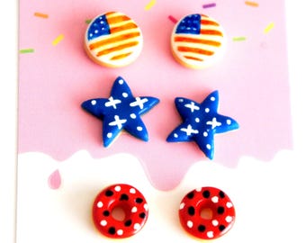 May include: Three pairs of stud earrings featuring miniature food-themed designs. The top pair are round cookies with American flag patterns. The middle pair are blue star-shaped cookies with white crosses. The bottom pair are red ring-shaped donuts.