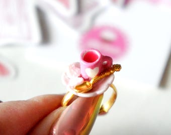 May include: A gold ring with a miniature pink teacup, saucer, and spoon. The teacup has a white heart design.