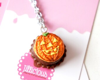 May include: A chocolate-covered pumpkin-shaped cake charm with a jack-o'-lantern face, hanging from a silver chain. The charm is a Halloween-themed accessory.