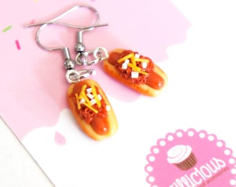 May include: A pair of novelty earrings featuring miniature hot dogs. Each earring has a silver-colored hook and a detailed hot dog charm with a bun, chili, and cheese topping. The earrings are set against a pink background.
