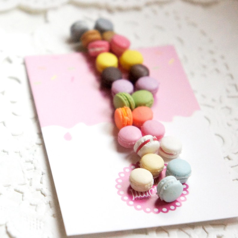 May include: A collection of colorful macarons arranged in a line on a pink background. The macarons are in various colors, including pink, blue, green, yellow, and orange.
