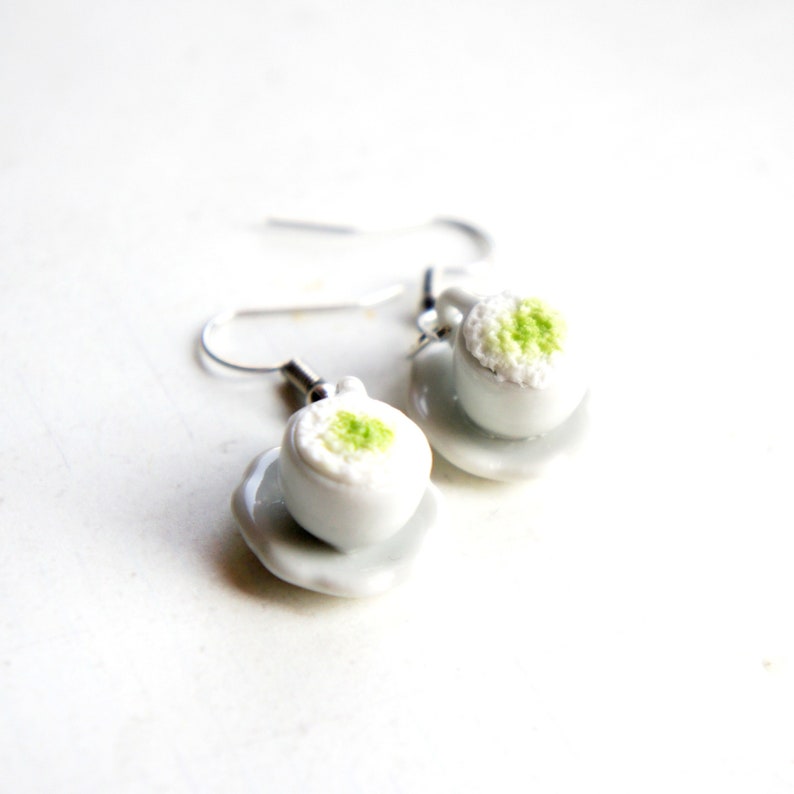 May include: A pair of silver dangle earrings featuring miniature white ceramic teacups with green tea inside. The teacups are on small white saucers.