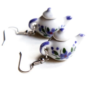 May include: A pair of silver dangle earrings with a white ceramic teapot charm. The teapot has a blue and purple floral design.