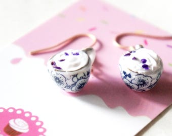 May include: A pair of gold dangle earrings featuring a miniature white ceramic teacup with a blue and white floral pattern and purple sprinkles on top.