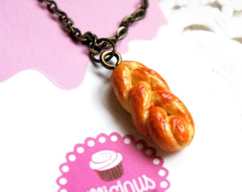 May include: A gold chain necklace with a brown braided bread charm. The charm is shaped like a loaf of bread and has a glossy finish.