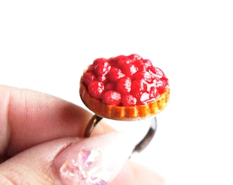 May include: A ring with a miniature cherry pie made of polymer clay. The pie is filled with red cherries and has a golden crust.