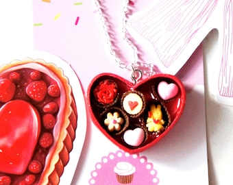 May include: A red heart-shaped box necklace with a silver chain. The box is filled with miniature chocolate candies, including a red rose, a heart, and a cupcake. The necklace is on a pink background with white polka dots and the text "jillicious charms and accessories".