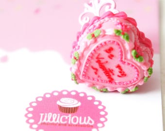 May include: A pink heart-shaped cake with white frosting and green accents. The cake is decorated with the words "Be my Valentine" in red frosting. The cake is sitting on a white surface with a pink and white logo that says "Jillicious" in pink.