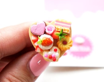 May include: A heart-shaped miniature food arrangement featuring a variety of desserts. The arrangement includes a pink heart, a rose, strawberries, and a small cake. The desserts are arranged on a light brown base, with a pink background.