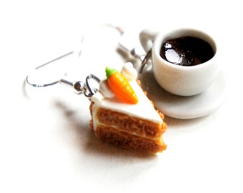 May include: A pair of dangle earrings featuring a miniature carrot cake slice with white frosting and a small orange carrot on top. The earrings are silver and have a hook closure. The cake slice is next to a miniature white coffee cup with a brown liquid inside.