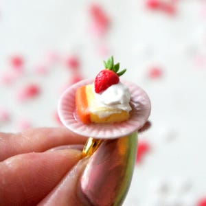 May include: A pink ring with a miniature cake and strawberry on a small pink plate. The cake is white with a yellow layer and a red layer. The strawberry is red with green leaves.