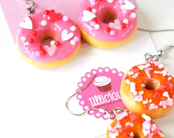 May include: Pink donut earrings with white, red, and pink sprinkles and heart-shaped accents. The earrings have a yellow base and silver hooks. The image also shows orange donut earrings with sprinkles and a pink tag with a cupcake illustration.