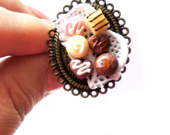 May include: An oval-shaped ring with a bronze-colored filigree frame. The ring features a miniature dessert arrangement, including pink, yellow, and brown candies on a white lace doily. The candies have white and brown decorative icing.
