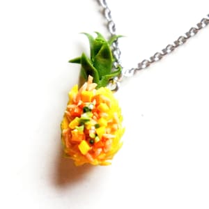 Puede incluir: Un collar con forma de piña con una cadena plateada. La piña está llena de arroz amarillo, verduras cortadas en dados de color naranja y rojo, y cebollas verdes.