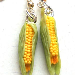 May include: A pair of earrings featuring miniature corn on the cob, with yellow kernels and green husks. The earrings are made of polymer clay and have silver hooks.