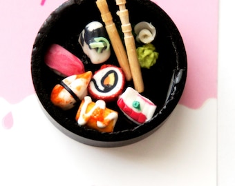 May include: Miniature sushi set in a black bowl with wooden chopsticks. The sushi includes various colorful pieces, such as nigiri, maki rolls, and wasabi. The background is a light pink with colorful sprinkles.