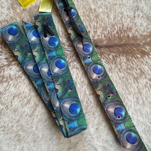 May include: A set of three fabric straps with a peacock feather design in shades of blue, green, and gold. One strap is tied with a black cord. A yellow tag with text is visible in the upper left corner.