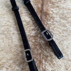 May include: Two black leather straps with silver-colored buckles. The straps have multiple adjustment holes and are designed for equestrian use. The background is a mottled brown and tan pattern.