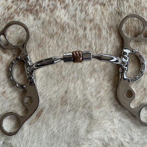 May include: A silver horse bit with a copper roller and a curved mouthpiece. The bit has two rings on each side, one larger than the other.