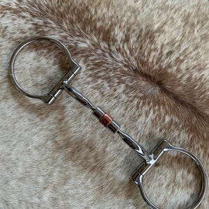 May include: A silver-colored metal horse bit featuring two large circular rings on each end and a central mouthpiece with a copper-colored roller segment in the middle. The bit is constructed with a jointed design, connecting the rings to the bar.