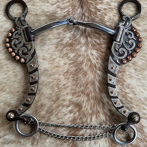 May include: A silver and copper horse bit with a brown leather mouthpiece and a chain attached. The bit has a floral design and is decorated with copper studs.
