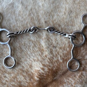 May include: A silver-colored horse bit with a twisted center and rounded rings. The bit is designed for equine use and is made of metal. The image is taken from a top-down perspective, showcasing the bit's intricate design.