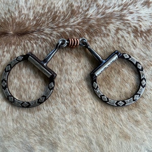May include: A silver and copper horse bit with decorative diamond patterns on the black rings. The bit is designed with a jointed mouthpiece and is displayed on a cowhide background.