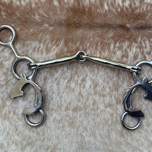 May include: A silver-colored horse bit with a curved mouthpiece and decorative horse head accents. The bit features rings and a central joint, designed for equine use. The background is a brown and tan mottled pattern.