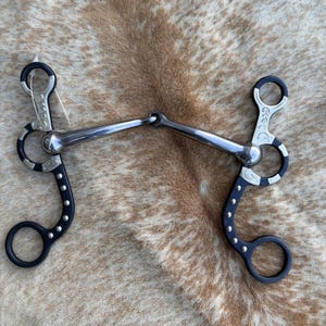 May include: A metal horse bit featuring a jointed mouthpiece and two black shanks. The shanks are decorated with silver-colored engravings and a series of small silver studs along the curved edges. The bit includes circular rings at the top and bottom.