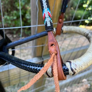 Op de afbeelding: Close-up van een paardenhoofdstel met een kralen frontriem. De frontriem heeft een patroon van witte, zwarte, rode en blauwe kralen. Het hoofdstel heeft bruine leren riemen en een zwart gevlochten touw. De teugels zijn wit en zwart.