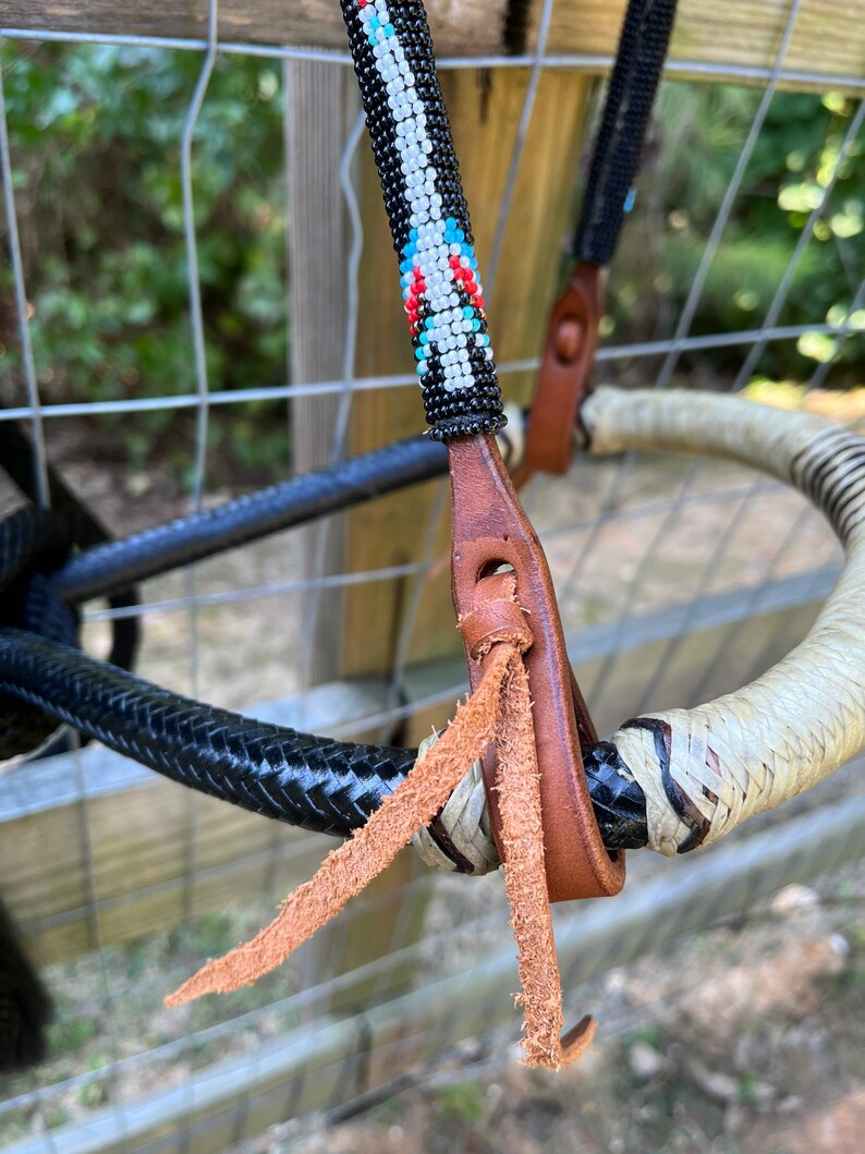 Op de afbeelding: Close-up van een paardenhoofdstel met een kralen frontriem. De frontriem heeft een patroon van witte, zwarte, rode en blauwe kralen. Het hoofdstel heeft bruine leren riemen en een zwart gevlochten touw. De teugels zijn wit en zwart.