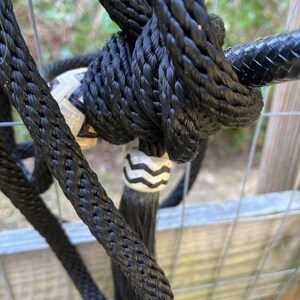 Op de afbeelding: Close-up van een zwart touwhalster met ingewikkelde knopen. Het halster heeft een decoratieve, patroon kraal en een lange, zwarte kwast. De achtergrond bestaat uit een metalen hek en wazig groen.