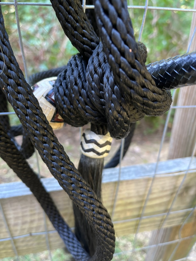Op de afbeelding: Close-up van een zwart touwhalster met ingewikkelde knopen. Het halster heeft een decoratieve, patroon kraal en een lange, zwarte kwast. De achtergrond bestaat uit een metalen hek en wazig groen.