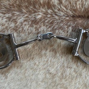 May include: A silver horse bit with a horseshoe and horse head design on each side. The bit is made of metal and has a central joint. The background is a brown and tan animal hide.