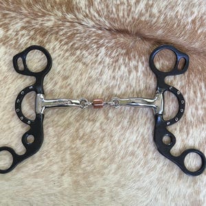 May include: A horse bit featuring a black metal shank with a curved design and silver-colored accents. The mouthpiece is made of polished silver-colored metal with a central copper-colored roller. The word SHOWMAN is engraved on the metal.
