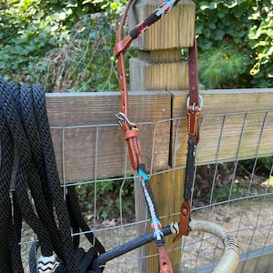 Op de afbeelding: Een paardenhoofdstel met een kralen voorhoofdsband en wangkappen. Het hoofdstel heeft een bruine leren kopstuk en een zwart touw met een decoratief uiteinde. De kralen secties hebben zwarte, rode, witte en turquoise kralen. Het bit is gewikkeld in een lichtgekleurd materiaal.