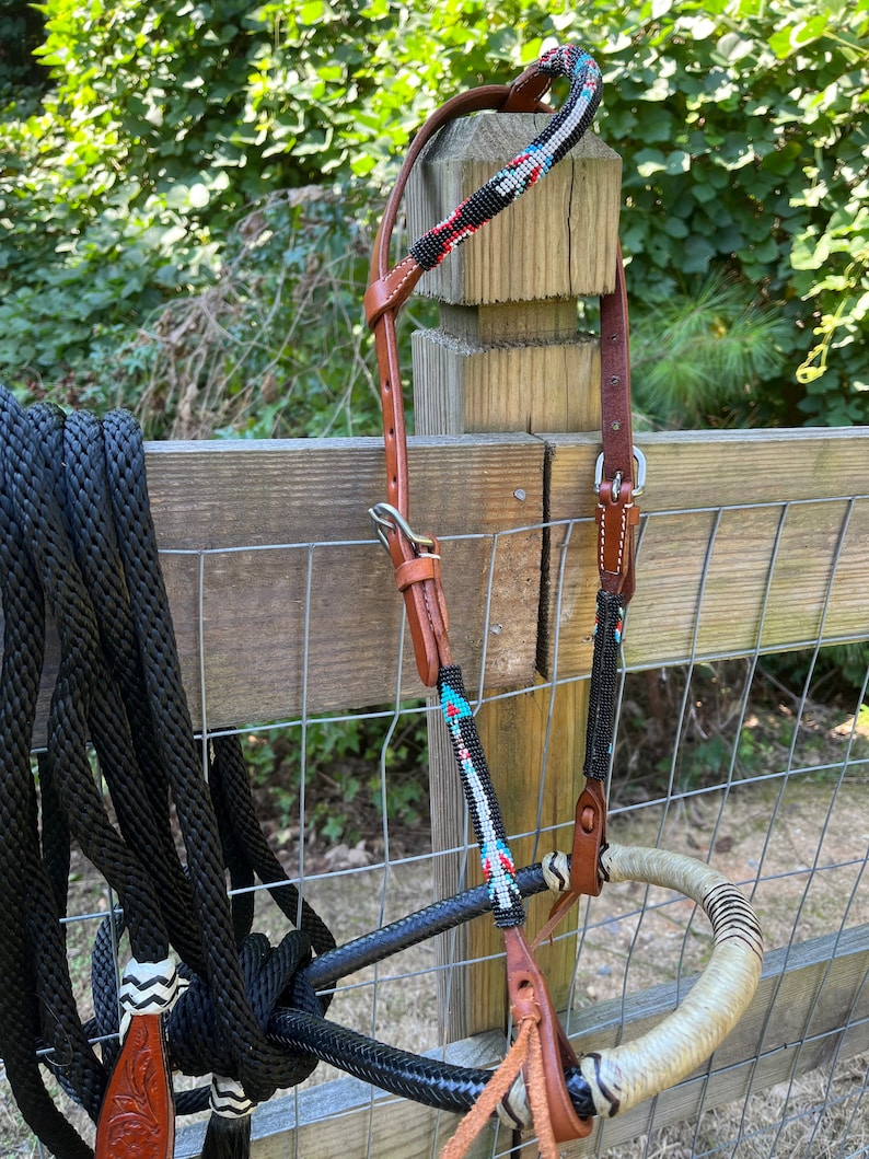 Op de afbeelding: Een paardenhoofdstel met een kralen voorhoofdsband en wangkappen. Het hoofdstel heeft een bruine leren kopstuk en een zwart touw met een decoratief uiteinde. De kralen secties hebben zwarte, rode, witte en turquoise kralen. Het bit is gewikkeld in een lichtgekleurd materiaal.