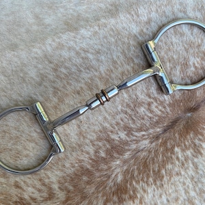 May include: A silver-colored horse bit with a D-ring design. The bit features a central mouthpiece with copper-colored rings and a curved, polished metal construction. The background is a mottled brown and tan surface.