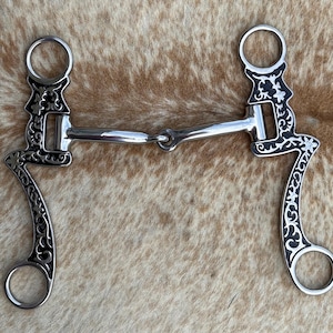 May include: A silver-colored metal horse bit featuring a jointed mouthpiece and two long, curved shanks. The shanks are decorated with intricate black and silver scrollwork patterns and include circular rings at the top and bottom ends.